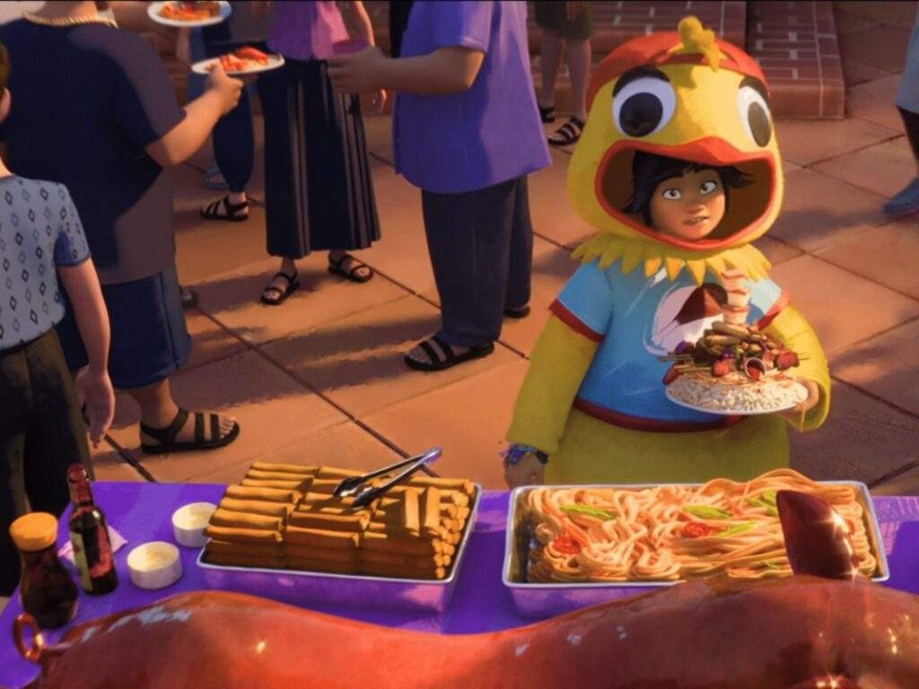 Jo, dressed in a bright yellow chicken costume, carries a plate piled with rice, barbecue, and an ice cream cone while standing in front of a buffet featuring lechon, pancit, and lumpia at a lively outdoor gathering.