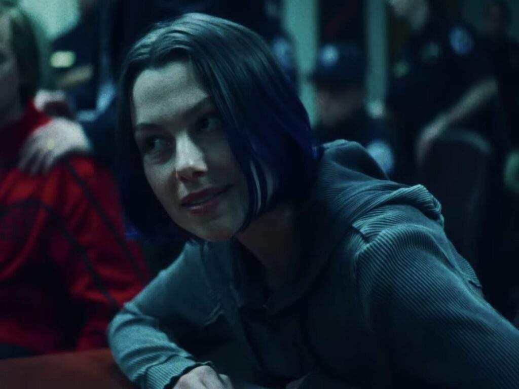 Phoebe Bridgers as decoy Del Harvey, wearing a gray collared shirt and leaning forward while looking to her left in a dimly lit, blue-toned room with several people in the background.