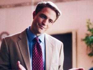 Robert Pattinson as Chris Hansen in Primetime (2026), wearing a beige suit, blue shirt, and red patterned tie while smiling and gesturing with his hands.