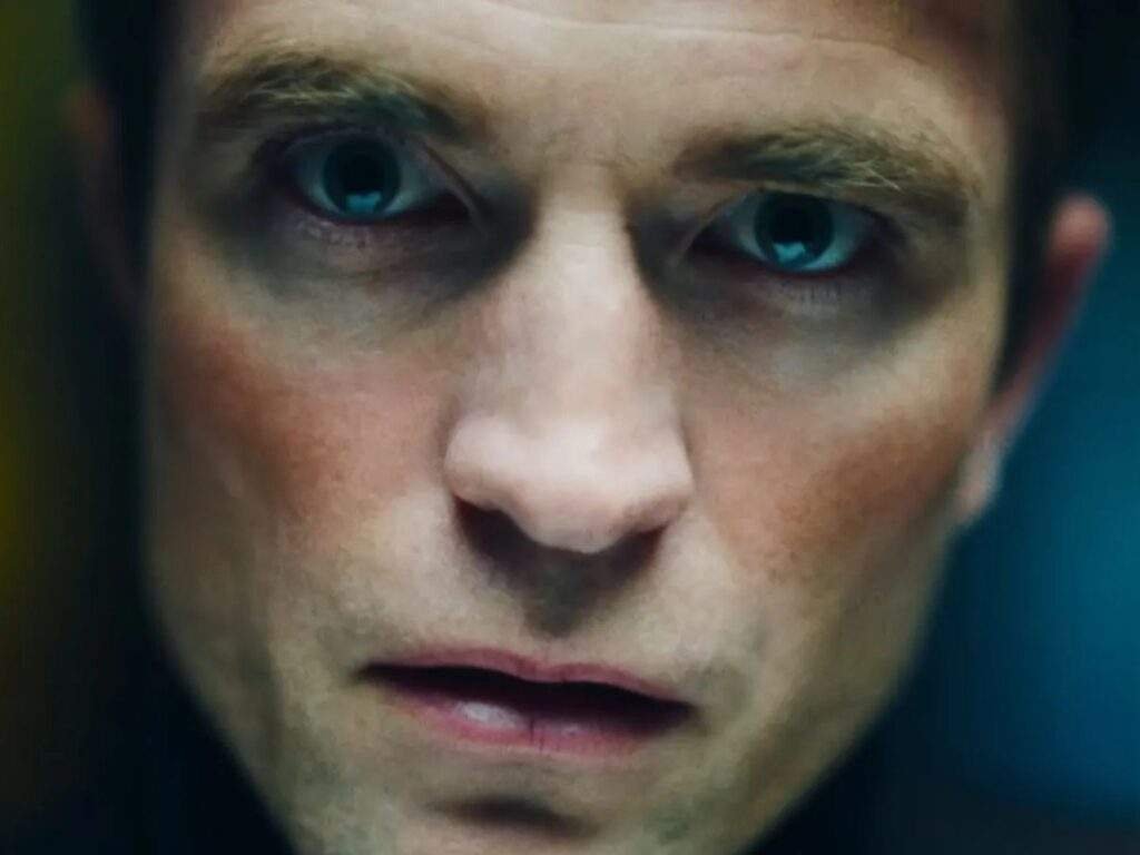 Extreme close-up of Robert Pattinson as Chris Hansen, staring intensely ahead with wide blue eyes, dark circles under his eyes, and slightly parted lips under dim, blue-green lighting. Primetime