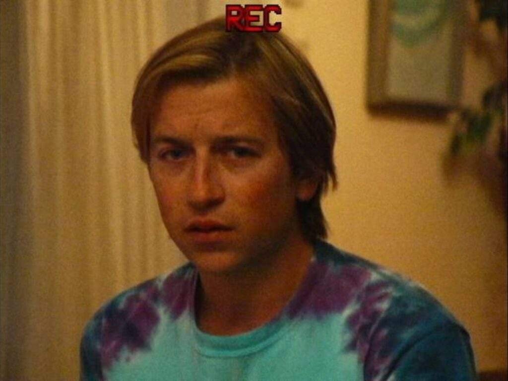 Skyler Gisondo as decoy Dan Plum, wearing a blue and purple tie-dye shirt and looking concerned in a dimly lit room, with a red "REC" recording indicator above his head. Primetime