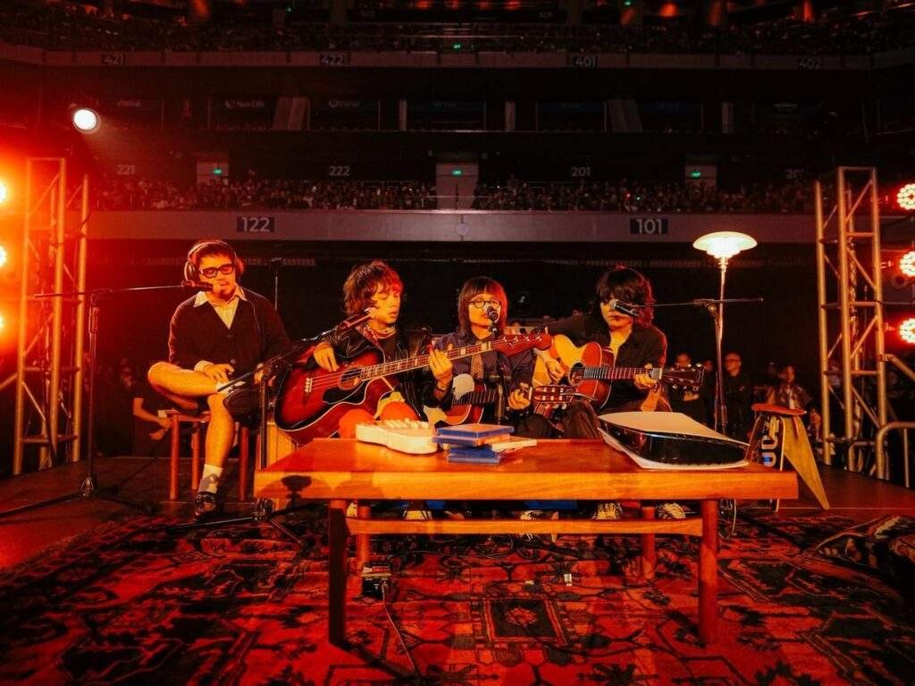 Filipino rock band IV of Spades performs an acoustic set at the SM Mall of Asia Arena, with all four members seated onstage playing guitars and singing into microphones under warm orange lighting, with the audience visible in the background.