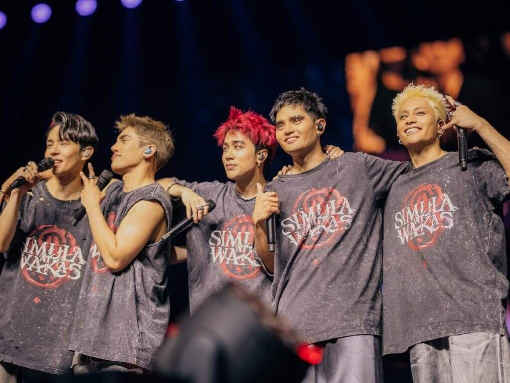 SB19 poses together onstage during their Simula at Wakas World Tour, with all five members wearing matching gray tour shirts, holding microphones, and putting their arms around one another under concert lighting.