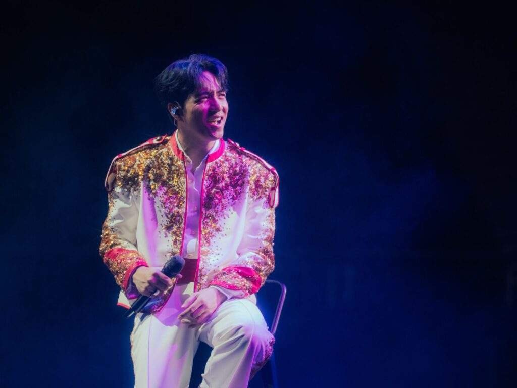 TJ Monterde smiles while seated on a stool during a concert, holding a microphone and wearing a white outfit with an ornate gold-sequined jacket under purple and blue stage lighting.